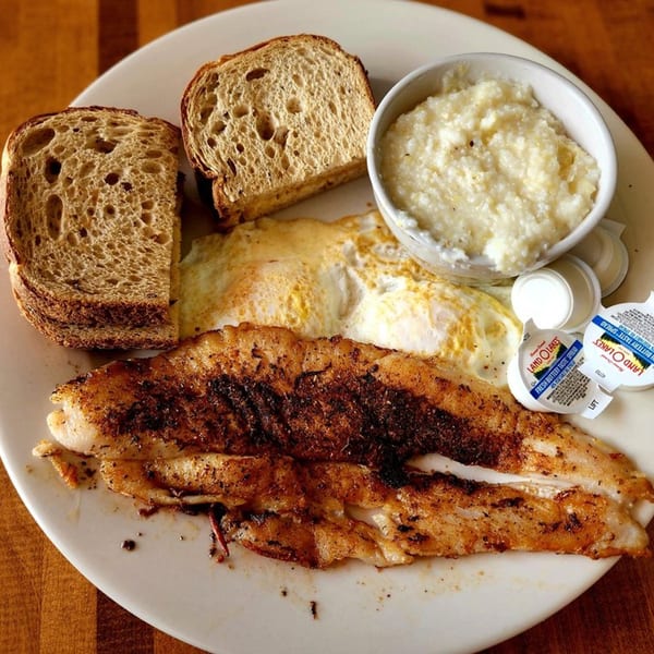 A breakfast plate with seasoned fish, fried eggs, grits, and two slices of toast with butter.