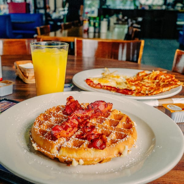 A waffle with bacon and powdered sugar, orange juice, and a breakfast plate with eggs and hash browns.