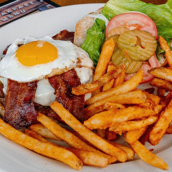 A breakfast burger with bacon, fried egg, lettuce, tomato, pickles, and a side of fries.